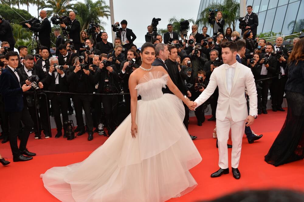 Priyanka Chopra wearing white tule strapless ballgown and diamond choker while holding Nick Jonas' hand as he wears an all-white suit with a bowtie and black shoes on the red carpet. Photo by Deposit Photos user Featureflash
